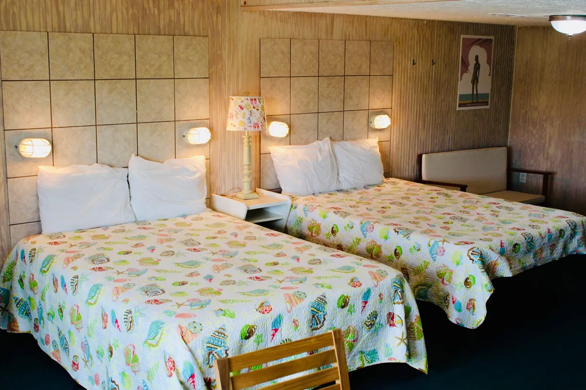 This image captures the heart of the studio's sleeping area, featuring two inviting beds with lively, colorful bedspreads. A central nightstand with a lamp offers a practical, cozy touch, while the wall art introduces personality and warmth. The setting is clearly designed for comfort, relaxation, and socializing, with ample space for friends or family to gather.
