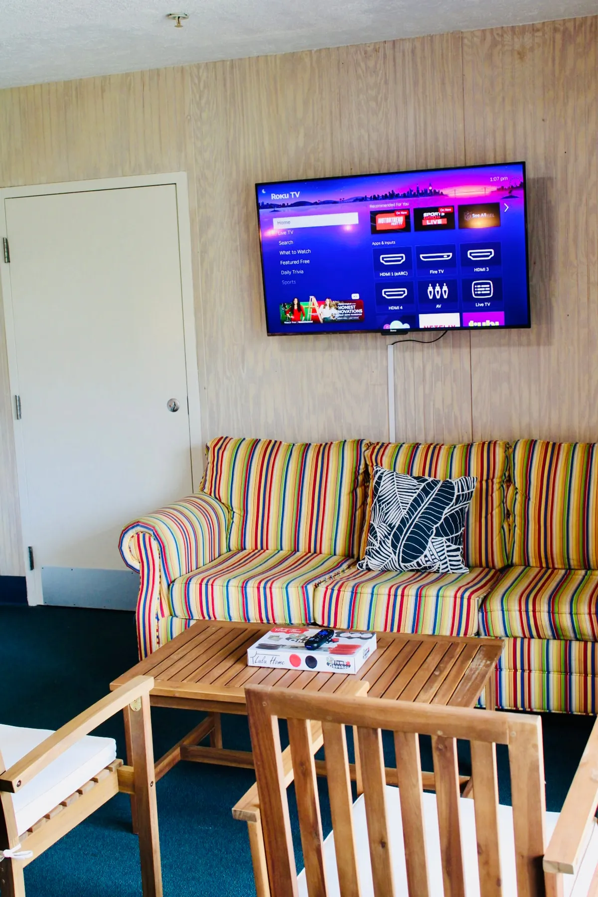 The inviting living area is centered around a striped couch and wooden coffee table, complemented by a Roku TV on the wall. The space is arranged for relaxation and shared activities, such as watching movies, playing games, or enjoying snacks as a group. The overall mood is casual and relaxed, with plenty of room for everyone, including pets, to stretch out.