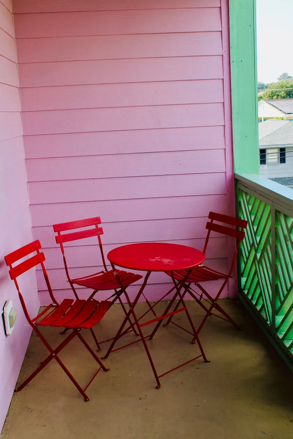 A playful exterior scene featuring bright red chairs and a matching table set against a pink wall with a green railing. The setup overlooks the outdoors, inviting guests to relax, sip coffee, or enjoy a book while taking in the vibrant atmosphere.