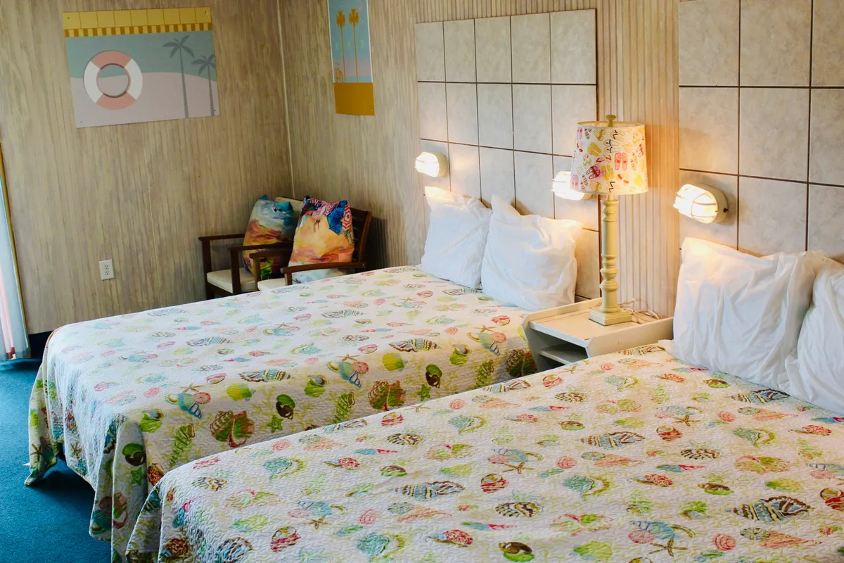 A cozy interior scene with two beds covered in colorful quilts, a decorative lamp, and art on the walls. The beds are bathed in natural light, evoking a welcoming space for relaxing or gathering for morning coffee or casual breakfasts.