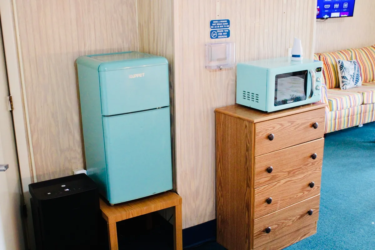 This image features a compact kitchenette with a vibrant turquoise full-size refrigerator, a microwave atop a wooden dresser, and a striped sofa nearby. The scene is warm and functional, illustrating both the cooking and relaxation spaces in one view.