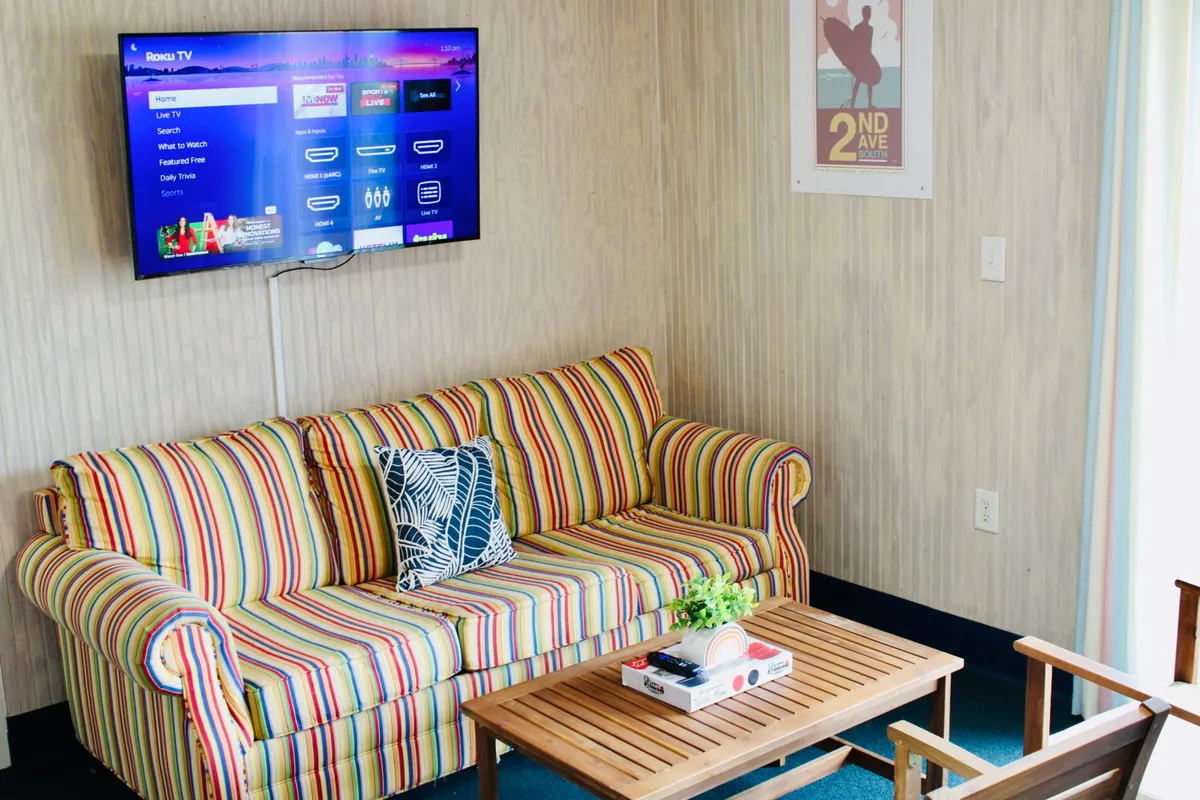 The image showcases the inviting interior of the studio, featuring a striped couch, a television, and a wooden coffee table, all arranged to create a cozy and social atmosphere. The space exudes comfort and functionality, perfect for relaxing after a day at the beach or gathering for movie nights and games with friends or family.